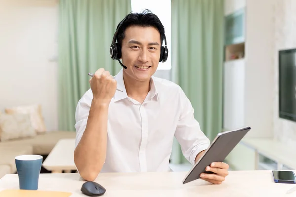 远程办公理念 亚洲男人带着耳机 高高兴兴地在家里用笔记本电脑和平板电脑参加一个视频会议 — 图库照片
