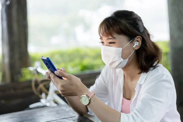 아시아의 여성은 보호용 마스크를 착용하고 이어폰 야외에서 행복하게 스마트폰을 — 스톡 사진