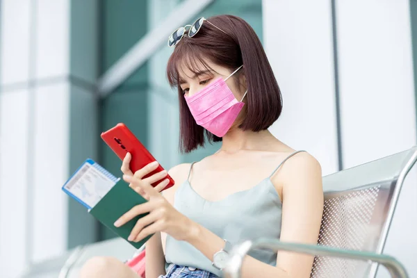 亚洲女性旅客使用带着面具的智能手机 在等候出发时坐在椅子上 — 图库照片