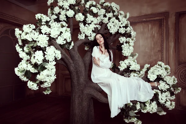 Krásná bruneta model sedí na kvetoucí dekorativní strom drží ruce u těla — Stock fotografie