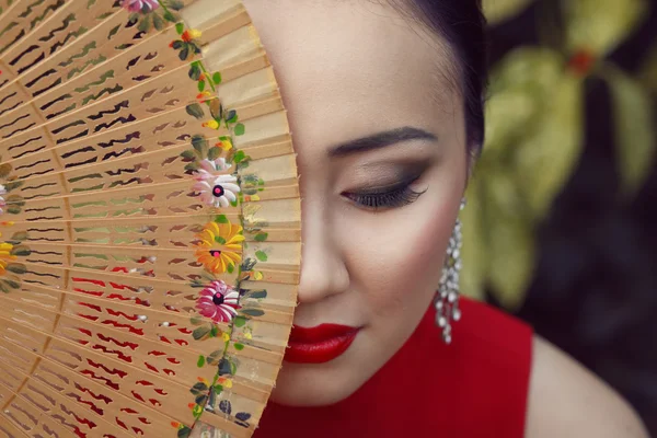 Linda asiática retrato feminino com vestido vermelho e lábios sentados no fundo floral verde segurando ventilador de madeira com padrão de flor perto de seu rosto — Fotografia de Stock