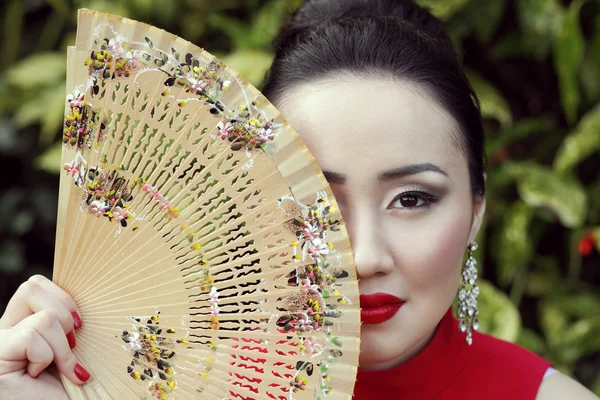 Horizontal portrait of japanese attractive female wearing red lipstick and dress and holding a bamboo fan with floral pattern in her hand