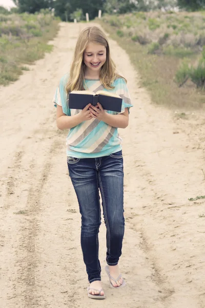 Glückliches Teenager-Mädchen liest ein Buch, während es eine unbefestigte Straße hinuntergeht — Stockfoto