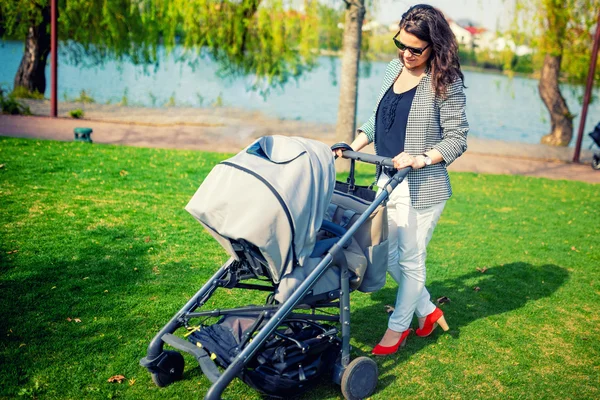 Mama elegantă zâmbește cu copilul în parc. Mama copil de mers pe jos cu cărucior sau cărucior pentru copii — Fotografie, imagine de stoc