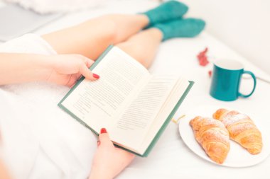 Seksi kadın kahvaltı ve kabarık sıcak çorap kruvasan ile yatakta kitap okumak Close-Up