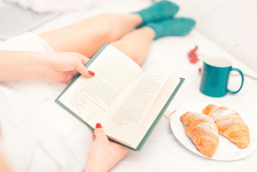 Close-up of sexy woman reading a book in bed with croissant as breakfast and fluffy warm socks