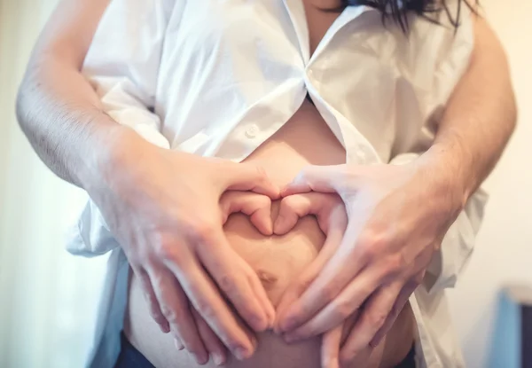 Concetto di maternità con coppia felice che si tiene per mano e crea un cuore sul pancione, sulla pancia incinta — Foto Stock