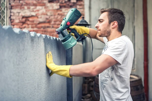 Homem usando luvas de proteção pintando uma parede cinza com pistola de tinta spray. Jovem trabalhador renovando casa — Fotografia de Stock