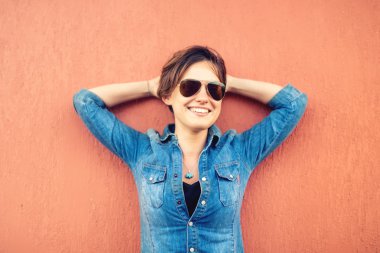 Trendy brunette girl, making face expressions, smiling and laughing against orange background, isolated. Modern lifestyle with beautiful hipster girl with sunglasses
