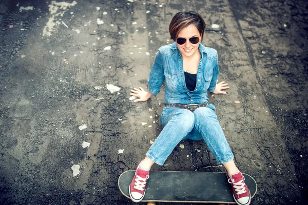 Modern girl with skateboard and sunglasses posing and smiling while sitting on terrace. Hipster girl wearing sunglasses and playing with skateboard — Zdjęcie stockowe