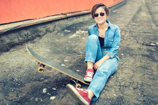 Chica morena con monopatín sentado, sonriendo a la cámara, con pantalones vaqueros y ropa moderna. Filtro de Instagram, concepto moderno de vida saludable —  Fotos de Stock