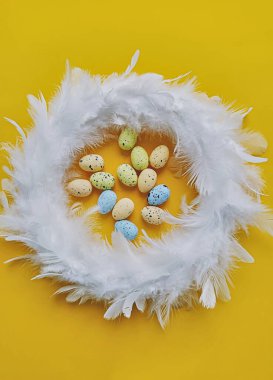 Sarı arka planda beyaz tüy çelengi ve küçük yapay yumurtalar, üst manzara. Paskalya dekorasyonu konsepti