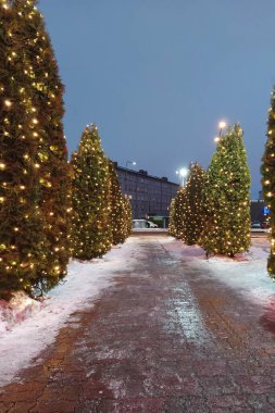 Karla kaplı yol aydınlanmış Noel ağaçları ve şehrin alacakaranlığında ılık festival ışıklarıyla kaplı.
