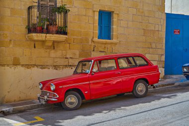 Eski güneşli kasabada taş binaların ve mavi kapıların yanına park edilmiş klasik kırmızı Ford station wagon Xghajra, Malta - 7 Haziran 2021
