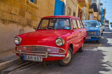 Taştan binaların ve mavi kapıların yanına park edilmiş kırmızı renkli Ford vagonun yakın görüntüsü: Xghajra, Malta - 7 Haziran 2021