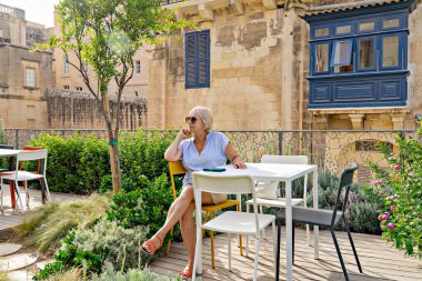 Kadın bahçedeki teras kafede oturuyor çiçekler ve yeşillikler arasında, tarihi taş binalar ve sıcak güneş ışığında mavi balkon.