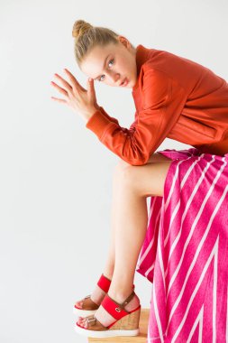 Kız dirsekleri dizlerine dayanmış şekilde oturur. Avuçlar katlanmış ve kafaya yakın. Elbiseler - turuncu ceket, çizgili etek ve yüksek kama sandaletler..