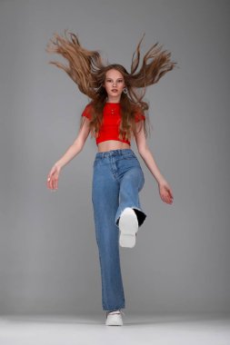 Stüdyoda poz veren yetişkin bir kız. Giysiler - kot pantolon, turuncu bluz ve spor ayakkabılar. Bacağıyla bir salıncak yapıyor, adımlarla. Saçları uçuyor, hareket ediyor..