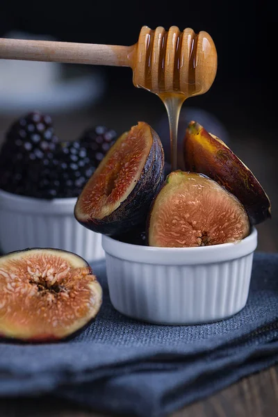 Honey pouring of fresh cuted organic figs and blackberry on white cupcake baking dishes on rustic wooden background with dark blue napkin. Low key with selective focus.