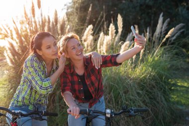 İki güzel beyaz kız akıllı telefonuyla selfie çekiyorlar. En iyi arkadaşlar bisiklet sürmenin tadını çıkarıyorlar. Güneşli yaz akşamı.