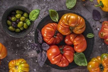 Whole and sliced organic tomatoes Raf Coeur De Boeuf. Raw fresh tomatos with sea salt and basil and olives in plates on rustic dark metal texture background
