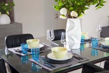 Modern evin siyah yemek masasında mavi ve yeşil tabak seti ve bir buket çiçek..