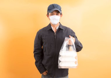Korumalı maskeli Asyalı kurye, sarı arka planda yemek kutusu tutuyor. Coronavirus konsepti 