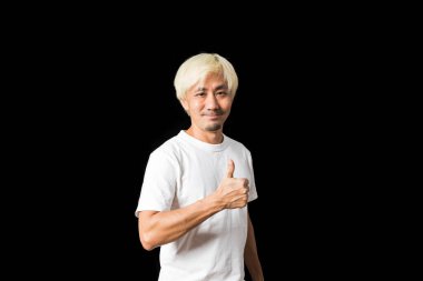 Orta yaşlı beyaz saçlı Asyalı adam siyah arka planda baş parmağını gösteriyor.   