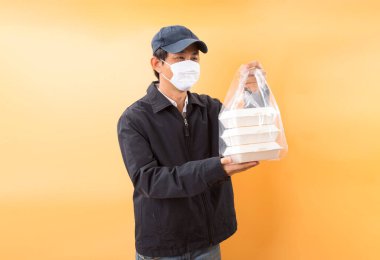 Korumalı maskeli Asyalı kurye, sarı arka planda yemek kutusu tutuyor. Coronavirus konsepti 