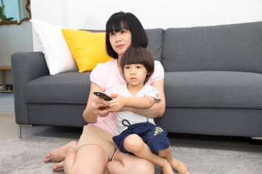 Asyalı anne ve oğul çift evdeki kanepede televizyon izliyor, anne oğluna uzaktan kumandayı nasıl kullanacağını öğretiyor..