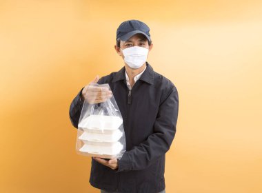 Korumalı maskeli Asyalı kurye, sarı arka planda yemek kutusu tutuyor. Coronavirus konsepti 