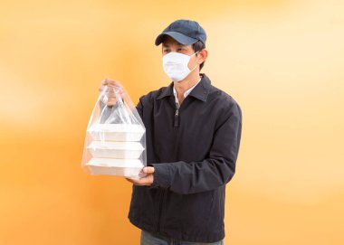 Korumalı maskeli Asyalı kurye, sarı arka planda yemek kutusu tutuyor. Coronavirus konsepti 