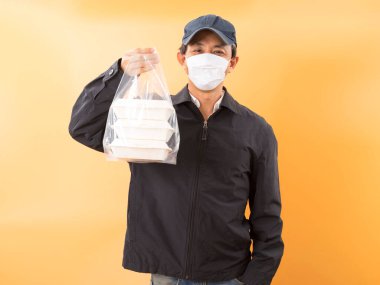 Korumalı maskeli Asyalı kurye, sarı arka planda yemek kutusu tutuyor. Coronavirus konsepti 