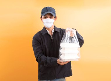 Korumalı maskeli Asyalı kurye, sarı arka planda yemek kutusu tutuyor. Coronavirus konsepti 