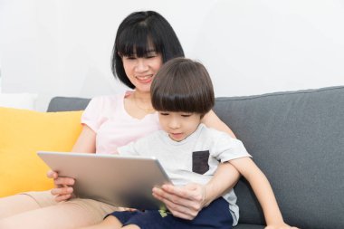 Mutlu Asyalı anne ve küçük tatlı oğlu karantina sırasında kanepede dinlenirken dijital tablette video izliyor ve izliyorlar..