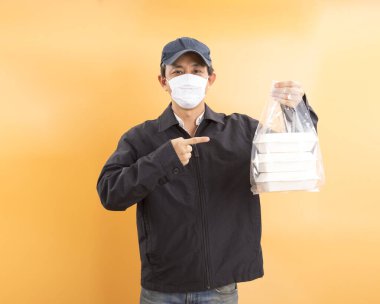 Korumalı maskeli Asyalı kurye, sarı arka planda yemek kutusu tutuyor. Coronavirus konsepti 