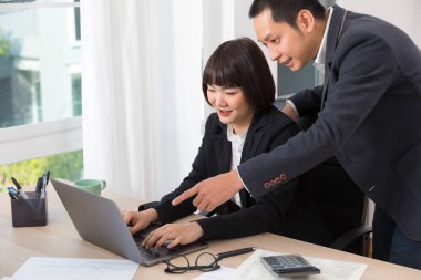 Asyalı işadamı ya da patron gülümseyip bilgisayarı işaret ederken kadın çalışan ve daktilo yazan.