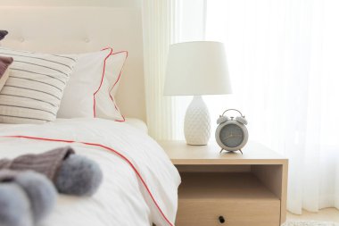 Masanın üzerinde lamba ve çalar saat, beyaz rahat yatağın yanında.