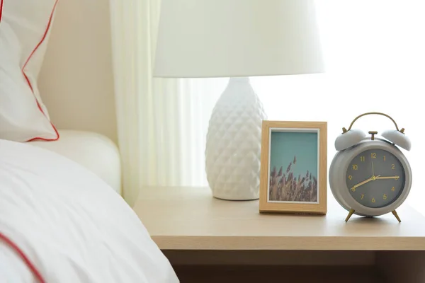 Komodinin üzerinde beyaz lamba ve çalar saat var.