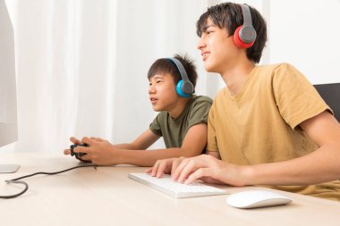 Kulaklıklı iki genç çocuk evde oyun oynamak için bilgisayar kullanıyor. Mutlu Asyalı çocuk evde online oyun oynuyor..
