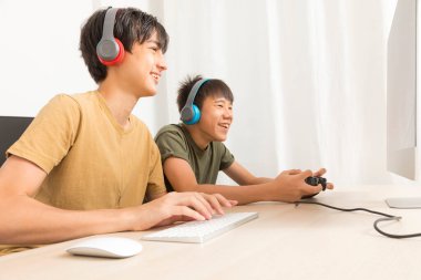 Kulaklıklı iki genç çocuk evde oyun oynamak için bilgisayar kullanıyor. Mutlu Asyalı çocuk evde online oyun oynuyor..