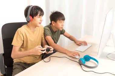Kulaklıklı iki genç çocuk evde oyun oynamak için bilgisayar kullanıyor. Mutlu Asyalı çocuk evde online oyun oynuyor..