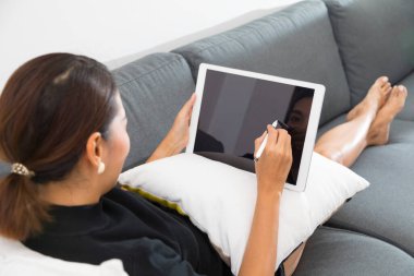 Genç Asyalı kadın kanepeye uzanıp evdeki dijital tablete bakıyor..