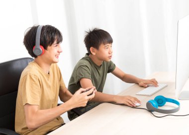 Kulaklıklı iki genç çocuk evde oyun oynamak için bilgisayar kullanıyor. Mutlu Asyalı çocuk evde online oyun oynuyor..