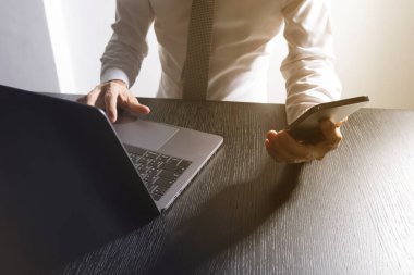 Beyaz gömlekli, akıllı telefonuna bakıp, masanın üstünde dizüstü bilgisayar kullanan iyi giyimli bir adamın ellerini yakın plan çek..