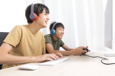 Kulaklıklı iki genç çocuk evde oyun oynamak için bilgisayar kullanıyor. Mutlu Asyalı çocuk evde online oyun oynuyor..