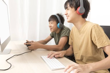 Kulaklıklı iki genç çocuk evde oyun oynamak için bilgisayar kullanıyor. Mutlu Asyalı çocuk evde online oyun oynuyor..