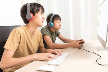 Kulaklıklı iki genç çocuk evde oyun oynamak için bilgisayar kullanıyor. Mutlu Asyalı çocuk evde online oyun oynuyor..
