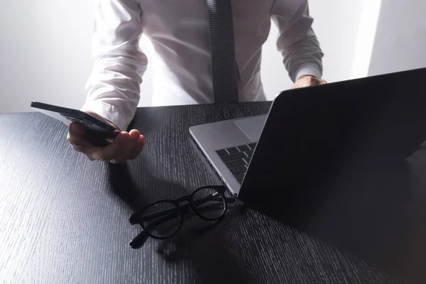 一位穿着白衬衫的男子的手部特写 他看着智能手机 在桌子上使用笔记本电脑 — 图库照片