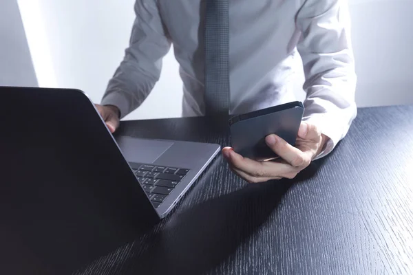 一位穿着白衬衫的男子的手部特写 他看着智能手机 在桌子上使用笔记本电脑 — 图库照片